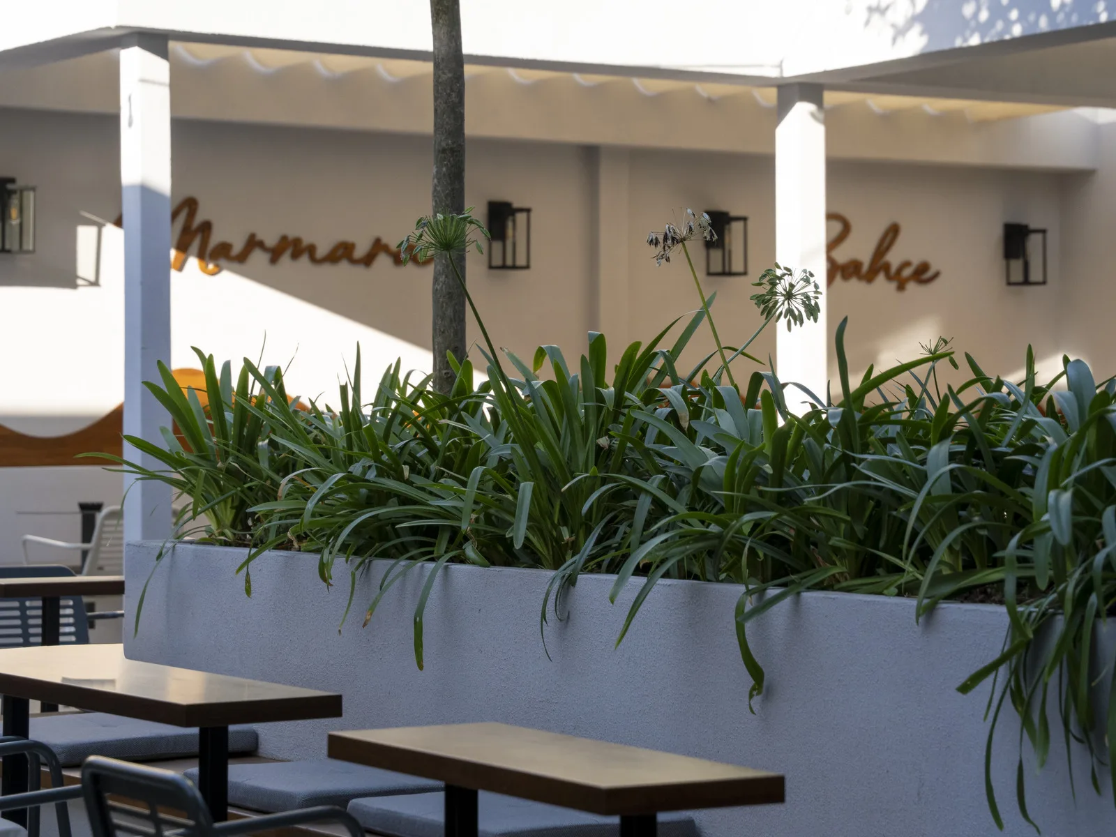 Ahşap masaların ardında bitkili uzun saksı, arkada pergola ve bakır renkli harfli duvar
