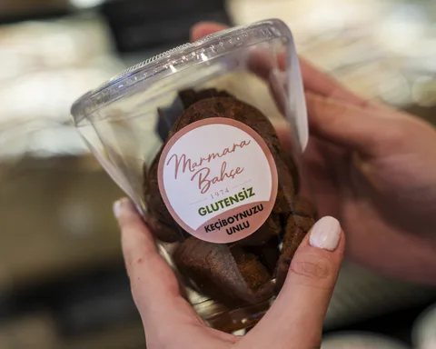 Şeffaf kapta koyu renkli kurabiyeler; Marmara Bahçe etiketinde Glutensiz ve Keçiboynuzu Unlu yazıyor.